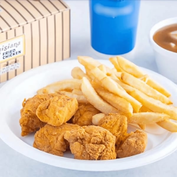 A white plate holds crispy fried chicken pieces and golden french fries, with a drink and sauce in the background.