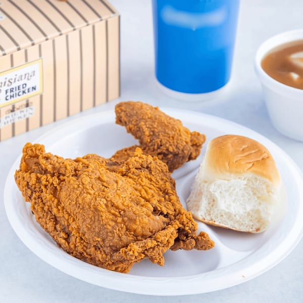 Two pieces of fried chicken and a dinner roll on a white plate with a drink and gravy.